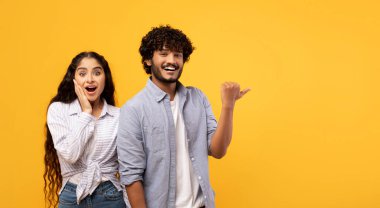 Omg, check this. Emotional indian guy pointing finger aside at free space over yellow background, surprised lady looking at camera, panorama. Couple demonstrating place for your text