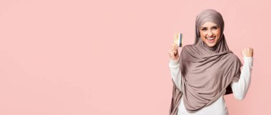 I made it. Overjoyed muslim girl in headscarf celebrating success with gold credit card, raising fist and looking at camera, pink background