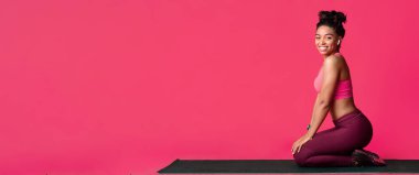 Genç atletik afro kız sportif sağlıklı yaşam tarzını seviyor yoga minderinde oturuyor kulaklıklarla müzik dinliyor ve kırmızı arka planda kameraya gülümsüyor.