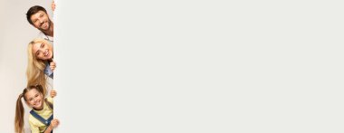 İki yetişkin ve çocuktan oluşan neşeli bir aile sevinçle boş bir duvarın arkasına bakıyor. Anne babalar geniş gülümseme sergilerken, kız da keyif veriyor, neşeli bir atmosfer yaratıyor, boşluğu kopyalıyor.