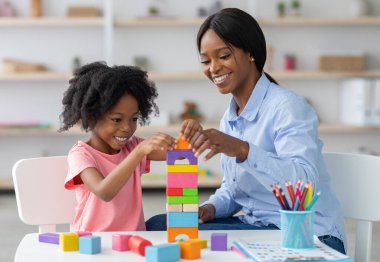 Sevimli siyah çocuk kıvırcık küçük kız ve çocuk gelişimi uzmanı çekici genç kadın renkli ahşap tuğlalarla oynuyor, masada oturuyor ve birlikte piramit yapıyor, gülümsüyor.