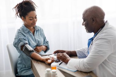 Gestational Hypertension. Mature Male Medical Worker Measuring Arterial Blood Pressure Of Pregnant Black Woman Using Cuff, Patient Having Problems With Tension, Sitting At Table. Health Care Concept