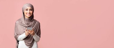 Müslümanlar için en iyi uygulamalar. Türbanlı gülümseyen İslamcı kız elinde akıllı bir telefonla pembe stüdyonun arka planındaki kameraya bakıyor.