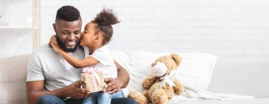 Afrikalı Amerikalı bir baba rahat bir koltukta oturur, küçük bir hediye tutar ve kızı onu neşeyle kucaklar. Küçük kız sevgisini bir öpücükle ifade ediyor.