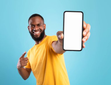 Rahat giyinen heyecanlı siyah adam cep telefonuna işaret ediyor mavi ekranlı arka plan, web sitesi ya da mobil uygulama tasarımı için alan. Cep telefonu ekran modeli. Seçici odak