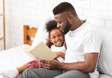 Mutlu akşamlar. Küçük kızına peri masalı okuyan Afrikalı-Amerikalı adam, yatak odasında, boş alanda oturuyor