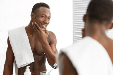 Erkek Cilt Bakımı. İçerideki banyoda aynanın yanında duran cilt bakımı yapan neşeli Afrikalı adam pürüzsüz yüze dokunuyor. Erkek Güzelliği ve Kişisel Bakım Rutini. Seçici Odaklanma