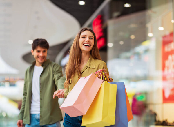 Вместе ходим по магазинам. Радостная жена таскает сумки с покупками, держа мужа за руку и смеясь наслаждаясь продажами в современном торговом центре. Торговля и шопоголизм. Селективный фокус