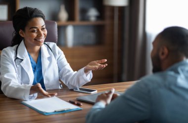 Friendly attractive african american young woman doctor having conversation with male patient, general practitioner consulting young man, gesturing and smiling, giving details, modern clinic interior
