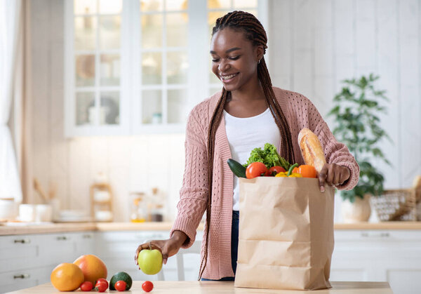 Black Smiling Lady Unpacking Paper Bag With Fresh Organic Vegetables And Fruits After Grocery Shopping, Happy African American Woman Standing In Cozy Kitchen Interior At Home, Copy Space