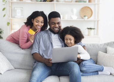 Çevrimiçi Alışveriş Konsepti. Küçük kızı olan mutlu bir Afro-Amerikan ailesi internetten mal almak için evde dizüstü bilgisayar ve kredi kartı kullanıyor.