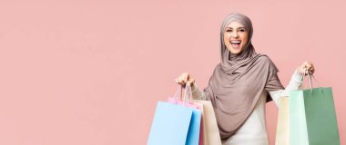 Büyük satışlar. Tesettürlü neşeli müslüman kız bir sürü parlak alışveriş torbası tutuyor başarılı alışverişin ardından mutlu oluyor, pembe arka planda boş alan ile poz veriyor.