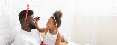 Kara baba ve kızı aydınlık bir yatak odasında neşeli bir an yaşıyorlar. Taç takan küçük kız, babasının yüzüne şakayla makyaj yapıyor ve yatakta oturuyor.