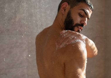 Cilt bakım konsepti. Genç kaslı adam ferahlatıcı bir duş alıyor. Vücudunu, sırtını ve çıplak omuzlarını sabun köpüğüyle yıkıyor. Sıcak su damlalarının altında banyoda duran kendinden emin yakışıklı adam.