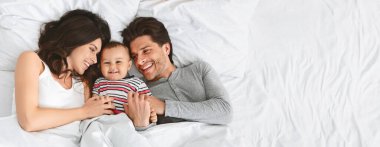 Yatakta birbirlerine sarılıp, gülümseyip gülerken neşeli bir aile bağı. Ebeveynler, küçük çocuklarıyla şakalaşarak sıcak ve sevgi dolu bir atmosfer yaratıyor ve uzayı kopyalıyorlar.