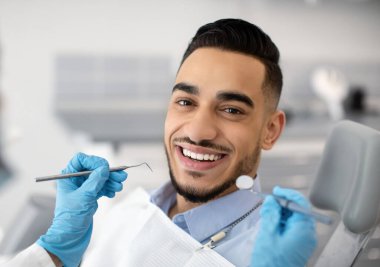 Modern klinikte dişçi koltuğunda oturan mutlu bir Arap 'ın portresi ve kameraya gülümseyen Orta Doğulu erkek hasta profesyonel stomatologla diş tedavisinden zevk alıyor.