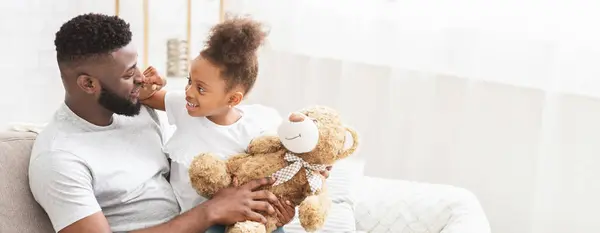 stock image African American father and his daughter share a delightful moment together in their cozy living room. The little girl holds a teddy bear while smiling at her dad, copy space