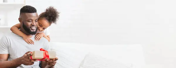 stock image African American father and his young daughter share a joyful moment as they unwrap a beautifully wrapped gift. The child eyes light up with excitement, while the father smiles warmly, copy space