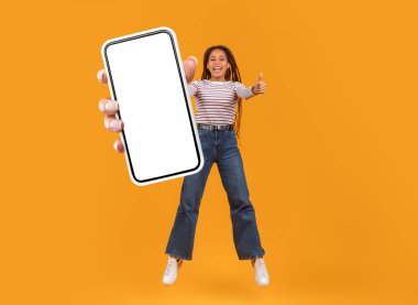 Uzun saçlı Afro-Amerikalı genç bir kadın, büyük bir akıllı telefonu tutarken, coşkuyla baş parmak kaldırıyor. Parlak sarı arka plan, o anın neşeli atmosferini arttırır..