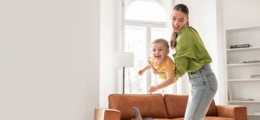 Bir anne, küçük oğlunu, aydınlık bir oturma odasında neşeyle döndürür. Konforlu alan kahverengi bir kanepe ve geniş pencereler içeriyor, aralarında neşeli, neşeli bir atmosfer yaratıyor..