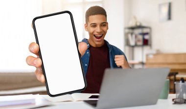 Genç siyahi bir adam, son zamanlardaki başarısını kutlamak için akıllı telefonunu kaldırırken sevinç ve heyecan ifade ediyor. O dağınık masasında oturmuş, hevesle dizüstü bilgisayarına bakıyor..