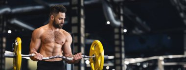 Kaslı bir adam, iyi donanımlı bir spor salonunda halterle biseps kıvırarak egzersiz yapar.