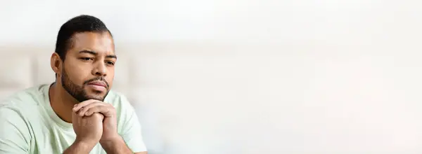 stock image African American man sits quietly in a light green shirt, deep in thought with his hands clasped. The soft lighting creates a calm atmosphere, suggesting a moment of introspection and contemplation.