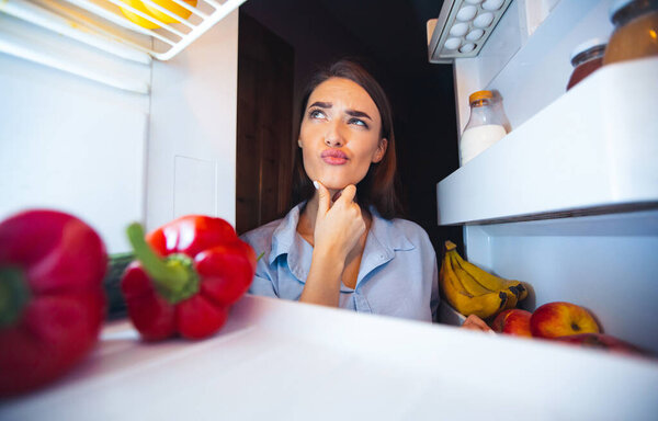 What should I eat. Young pensive woman thinking near opened fridge, view from inside