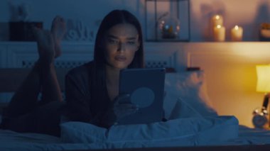 Global haberler. Ciddi bir kadın dijital tabletten haber okuyor, gece geç saatte yatakta yatıyor.