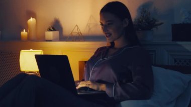 Gençlik iletişimi. Neşeli kız dizüstü bilgisayarında daktilo yazıyor, erkek arkadaşıyla gece geç saatlerde yatakta sohbet ediyor.