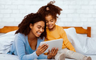 Aile Hafta Sonu. Mutlu Afrika kökenli Amerikalı anne ve kızı evde yatakta yatarken tablet bilgisayar kullanıyor..