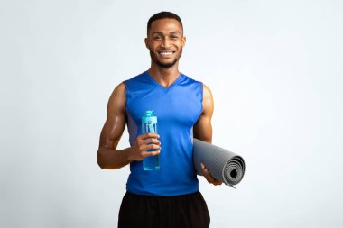 Fitness Konsepti. Elinde su şişesi ve yoga minderi tutan mutlu siyah adam, sarı duvarın üzerindeki kameraya gülümsüyor.