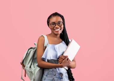Akademik eğitim. Sırt çantalı ve defterli genç siyah bir kadın pembe stüdyo arka planında kameraya gülümsüyor. Afro demetlerle okula başlamaya hazır genç bir üniversite öğrencisi.