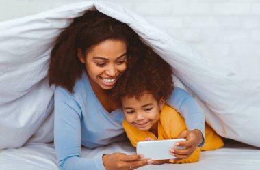 Afro-Amerikan anne ve kızı evde battaniyeyle örtülü olarak rahat bir yatakta uzanmış akıllı telefon kullanıyor. Seçici Odaklanma