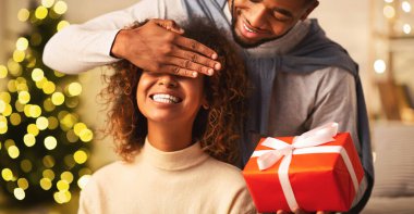 Mutlu afro adam kız arkadaşına Noel hediyesi veriyor, sürpriz yapıyor ve gözlerini kapatıyor.