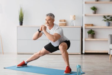 Ev serobiği. Olgun bir adam oturma odasında paspas üzerinde hareketler yapıyor spor kıyafetleri ve gülümseme giyerek ev içi fitness eğitiminden zevk alıyor.