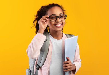 Education Concept. Portrait of positive smiling African American girl touching glasses holding textbooks, wearing backpack and looking at camera isolated on yellow studio background