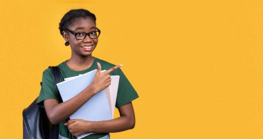 Sarı Stüdyo Arkaplanının üzerinde duran kitap ve defterlerle mesajlaşmak için boş uzayı işaret eden gülümseyen siyah kız öğrenci. Büyük Eğitim Teklif Reklamı