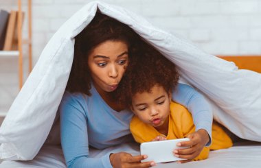 Afrikalı anne ve küçük kız, akıllı telefonlarıyla oyun oynuyorlar yatakta uzanıyorlar ve birlikte eğleniyorlar. Seçici Odaklanma