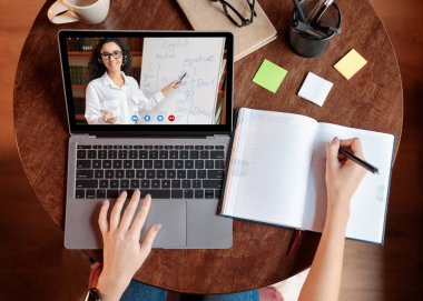 Uzaklık Eğitim Konsepti. Masada oturan, not defteri yazan, not alan, dizüstü bilgisayar kullanan, öğretmenle online ders alan üst düzey bir bayan öğrenci. Evden uzak çalışma, karantina