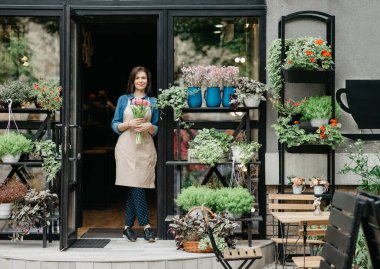 Startup, small business, eco restaurant outdoor and modern rustic flower shop. Smiling millennial beautiful female in apron holds bouquet of tulips for client, at front door of plants studio or cafe