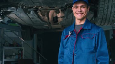 Dostça davranan, kameraya gülümseyen, el hareketi yapan, araba tamir atölyesini onaylayan bir otomobil tamircisi.