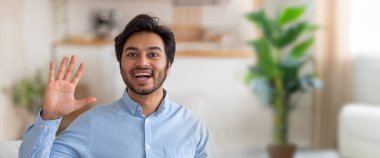 Neşeli genç Hintli bir adam, sıcak ve davetkar bir yaşam alanında geniş bir gülümsemeyle el sallıyor. Yakındaki bir bitkinin doğal ışığı ve yeşilliği ile dolu..