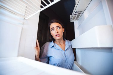 Şok olmuş genç bir kadın evdeki boş buzdolabına bakıyor, içeriden bakıyor.