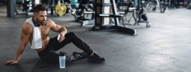 A young man sits on the gym floor, taking a break after exercising, with a towel around his neck and a water bottle nearby, copy space clipart