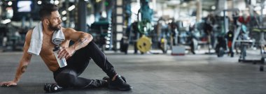 Formda bir adam spor salonunda, elinde bir su şişesi, antrenman kıyafetleri, fotokopi odasında rahatlar.