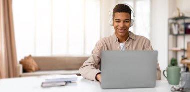 Genç siyah bir adam evde çevrimiçi bir öğrenim seansına katılıyor, kulaklık takıyor ve bilgisayarını kullanırken gülümsüyor. Parlak, doğal ışık rahat odayı doldurur..