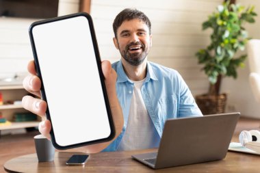 Bir adam elinde boş bir ekranla akıllı telefonu tutarken gülümsüyor, rahat bir ev ofisin masasında dizüstü bilgisayarı ve sıcak dekoruyla oturuyor..