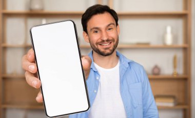 Bir adam parlak, davetkar bir odada duruyor, boş bir akıllı telefonu tutarken gülümsüyor. Arkaplanda raf dekoru var, sıcak bir atmosfer yaratıyor.