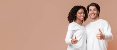 Çift onayı. Mutlu beyaz adam ve afro kız başparmaklarını kaldırıyor ve gülümsüyor, sarı arka planda birlikte poz veriyorlar, stüdyoda, boş alanda.